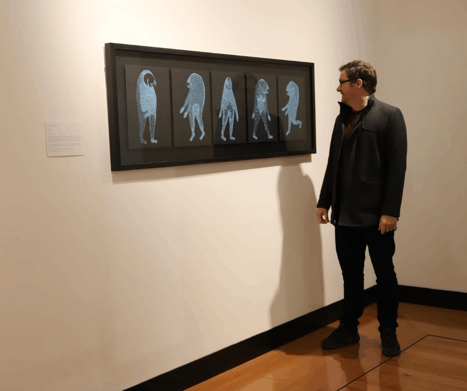 paranaple arts centre director, Geoff Dobson, looks at the 2014 tidal winning work Galaxias by Joel Crosswell. The artwork hangs on a wall. It is 5 pieces within a frame, each A4 size of black background, each piece has an alien like creature painted in blue, each creature has a fish shaped head.