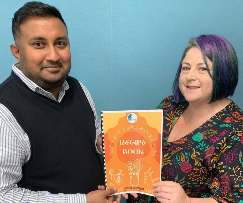 A man and a lady standing next to each other holding an orange recipe book to celebrate this year's Harmony Week.