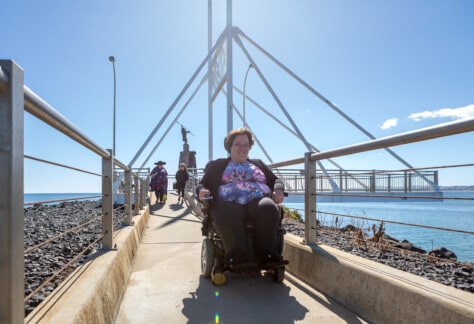 With a smile on her face Anne travels away from the Spirit of the Sea Sculpture in her wheelchair. She is wearing a pink and purple top with butterflies. Two people walk behind her, the sun is shinning, the sky and water are bright blue.