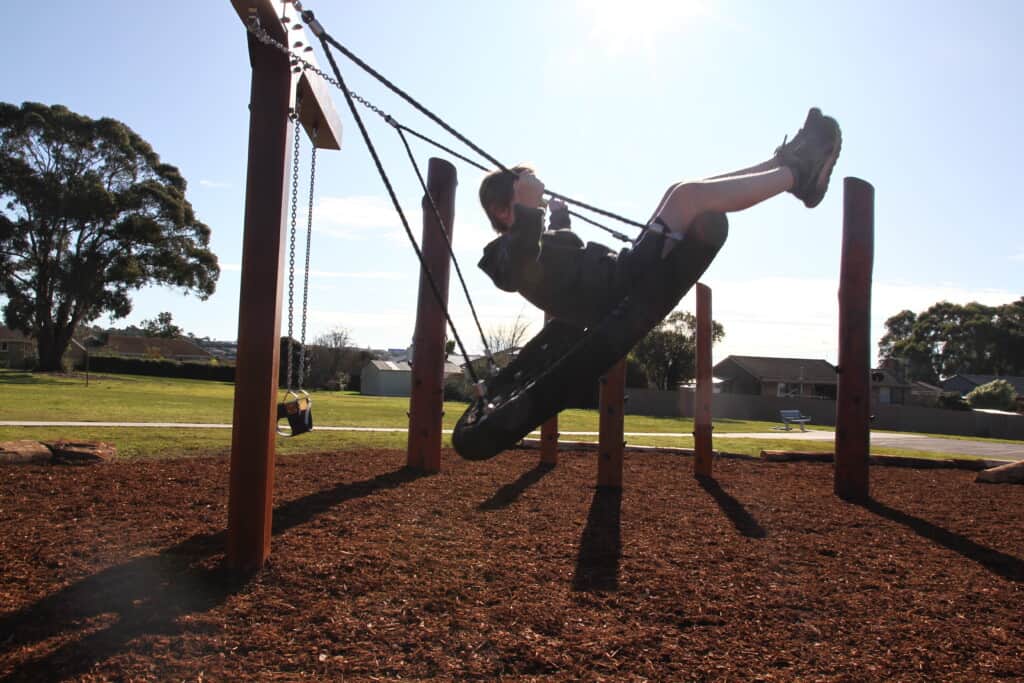 A child swings on a basket swing in a nature playground, the sun is shining brightly