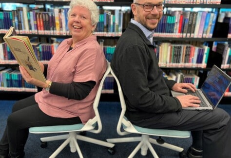 A woman and a man seated facing away from each other holding a book and a laptop with library books in the background.