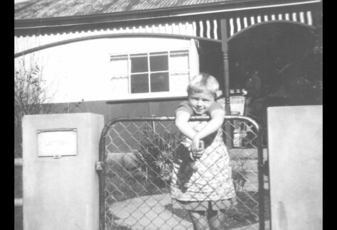 a child stands behind a fence, by the look of the house, fence and child's outfit it the 1950's