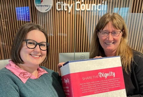 Two women standing next to each other holding a pink and white box which says "Share the Dignity". The box is for sanitary product donations to be made for the month of August.