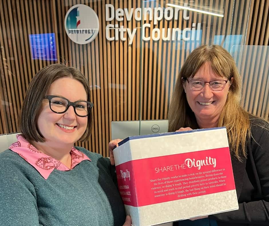 Two women standing next to each other holding a pink and white box which says "Share the Dignity". The box is for sanitary product donations to be made for the month of August.