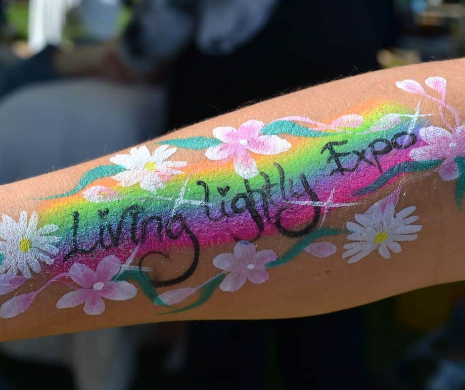 Image shows a painted arm. Theres a squiggly rainbow base, the words Living Lightly Expo in black, surrounded by pink and white flowers