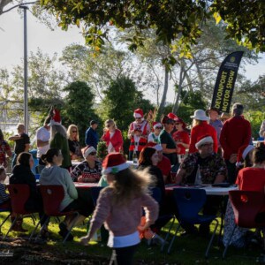 A group of people in Christmas attire sit at tables outside in a park setting