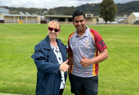 A woman and young man standing next to each other on a cricket pitch, giving the thumbs up, with the clubrooms in the backround.