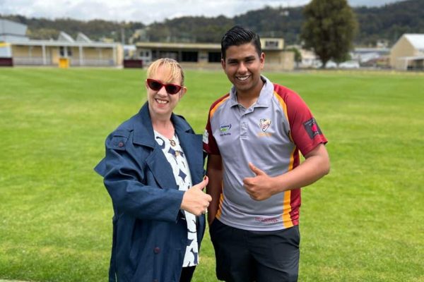 A woman and young man standing next to each other on a cricket pitch, giving the thumbs up, with the clubrooms in the backround.